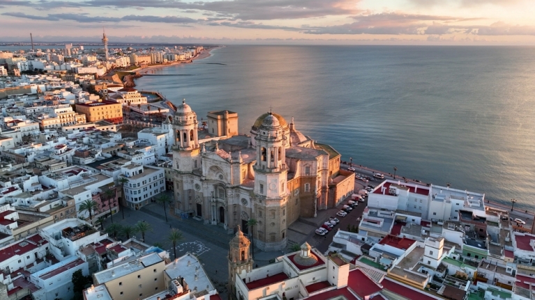 Una vista aérea de una iglesia y edificios circundantes en Cádiz al atardecer, con el océano al fondo